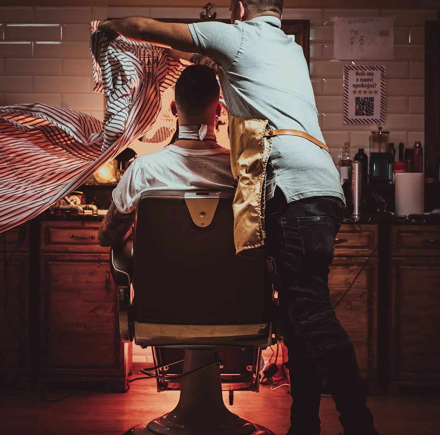 Barber prehadzuje strihací plášť cez zákazníka v kresle, v barbershope svieti červené svetlo
